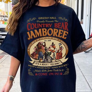 May include: Navy blue t-shirt featuring a vintage-style graphic for the "Country Bear Jamboree." The design includes cartoon bears playing instruments and the text "Grizzly Hall" and "Est. 1898." The shirt has short sleeves.