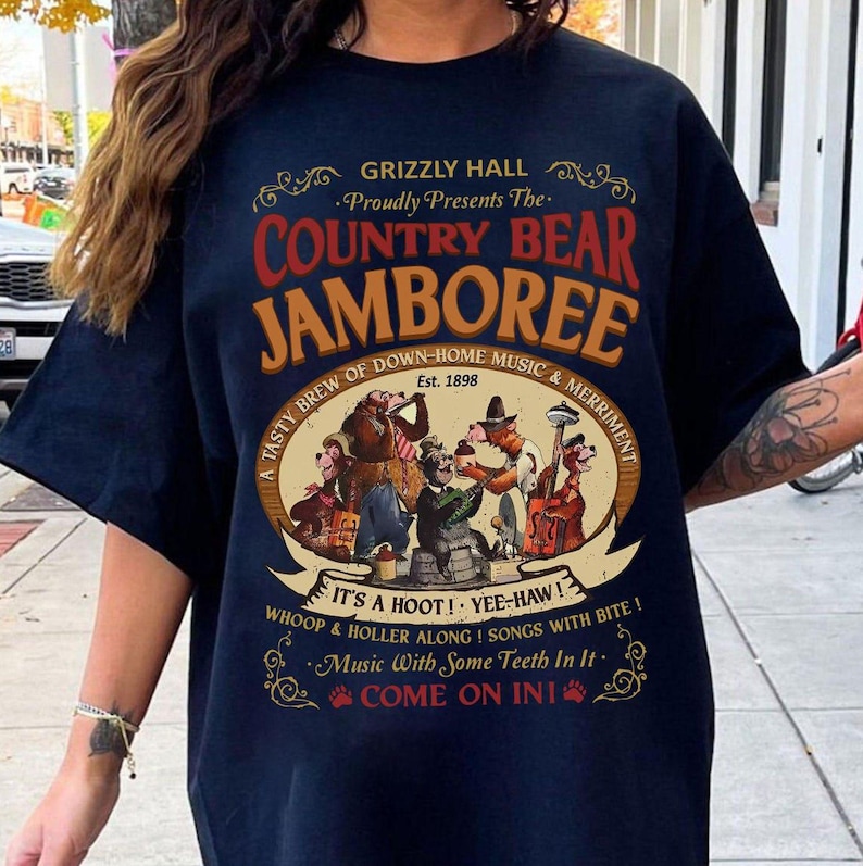 May include: Navy blue t-shirt featuring a vintage-style graphic for the "Country Bear Jamboree." The design includes cartoon bears playing instruments and the text "Grizzly Hall" and "Est. 1898." The shirt has short sleeves.