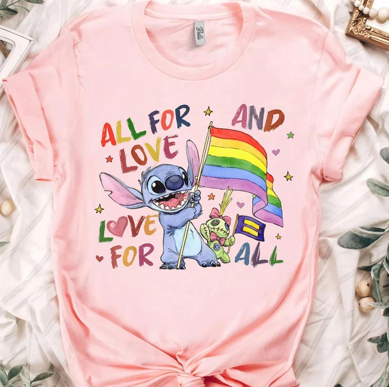 May include: Pink t-shirt featuring a cartoon character holding a rainbow flag. The shirt has the text "ALL FOR LOVE" and "LOVE FOR ALL" in a colorful design. The design includes stars and a second character.