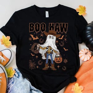 May include: Black t-shirt with a Halloween graphic. The design shows a cartoon character in a ghost costume, holding a pumpkin bucket and a lasso. The text "BOO HAW" is above the character in orange.