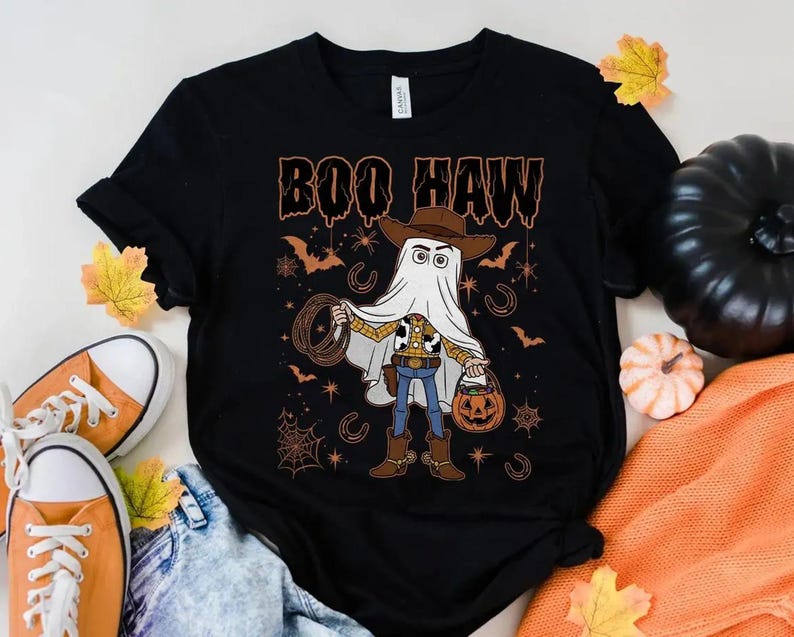 May include: Black t-shirt with a Halloween graphic. The design shows a cartoon character in a ghost costume, holding a pumpkin bucket and a lasso. The text "BOO HAW" is above the character in orange.