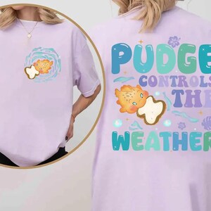 K&ouml;nnte beinhalten: Helllila T-Shirt mit einem Cartoon-Design eines Fisches und einer Wolke. Auf der R&uuml;ckseite des Shirts steht in einer farbenfrohen Retro-Schriftart "Pudge Controls The Weather". Das Shirt ist aus weichem Material.