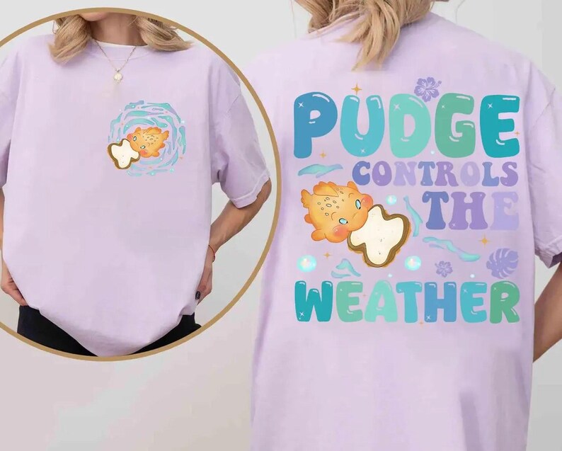 K&ouml;nnte beinhalten: Helllila T-Shirt mit einem Cartoon-Design eines Fisches und einer Wolke. Auf der R&uuml;ckseite des Shirts steht in einer farbenfrohen Retro-Schriftart "Pudge Controls The Weather". Das Shirt ist aus weichem Material.