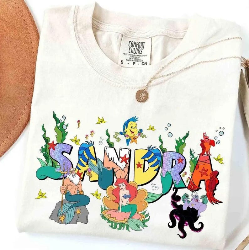 May include: Cream-colored t-shirt with the name "SANDRA" in colorful, stylized lettering. The design incorporates cartoon characters and aquatic elements, including mermaids, fish, and seaweed. A gold necklace with a pendant is draped on the shirt.