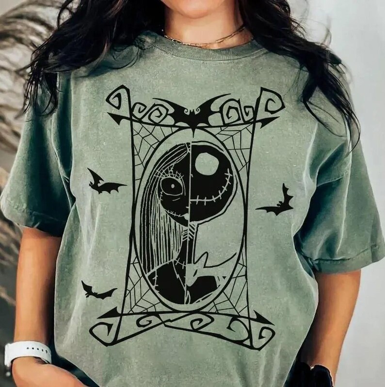 May include: Sage green t-shirt featuring a black graphic of a split-face character design within a decorative frame, with bat silhouettes. The design includes a half-face with stitches and a half-face with long hair. The shirt has short sleeves.