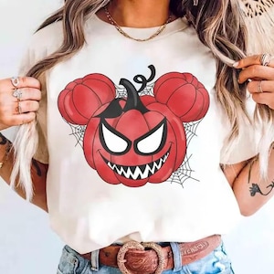 May include: A cream-colored short-sleeve t-shirt featuring a graphic print of a red pumpkin with a stem and two smaller round pumpkin ears, resembling a mouse silhouette. The pumpkin has a wide, toothy grin, white eyes, and black spiderwebs.