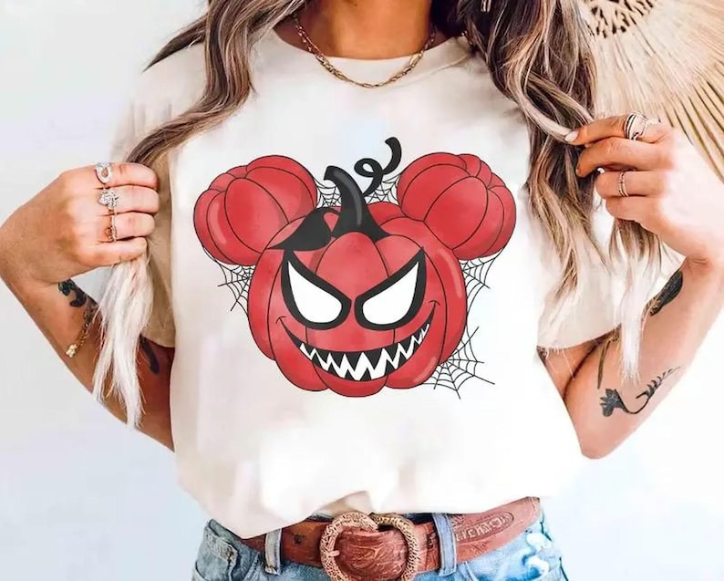May include: A cream-colored short-sleeve t-shirt featuring a graphic print of a red pumpkin with a stem and two smaller round pumpkin ears, resembling a mouse silhouette. The pumpkin has a wide, toothy grin, white eyes, and black spiderwebs.