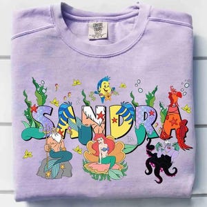 May include: A lilac-colored sweatshirt featuring the name "SANDRA" in a colorful, mermaid-themed design. The design includes illustrations of characters from "The Little Mermaid" movie, such as Ariel, Flounder, and Ursula.