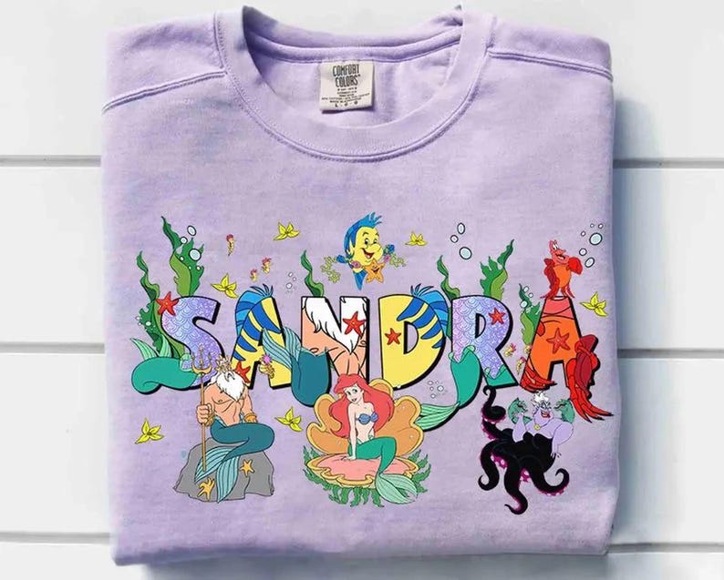 May include: A lilac-colored sweatshirt featuring the name "SANDRA" in a colorful, mermaid-themed design. The design includes illustrations of characters from "The Little Mermaid" movie, such as Ariel, Flounder, and Ursula.