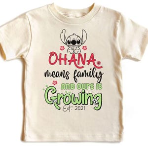 May include: Cream-colored t-shirt featuring a cartoon Stitch character above the words "Ohana means family and ours is growing Est. 2021" in red, black, and green. The design includes floral accents.