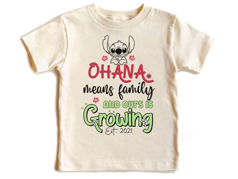 May include: Cream-colored t-shirt featuring a cartoon Stitch character above the words "Ohana means family and ours is growing Est. 2021" in red, black, and green. The design includes floral accents.