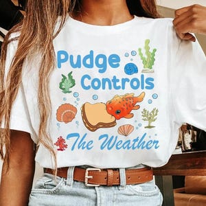 May include: White t-shirt with a colorful graphic design. The design features the text "Pudge Controls The Weather" in blue, along with illustrations of a fish, seashells, and seaweed.
