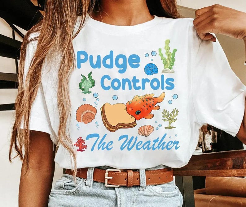 May include: White t-shirt with a colorful graphic design. The design features the text "Pudge Controls The Weather" in blue, along with illustrations of a fish, seashells, and seaweed.