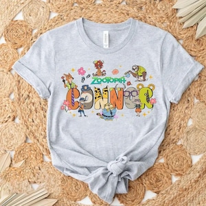 Puede incluir: Camiseta gris claro con la palabra "Connor" en letras coloridas estilo caricatura, y la palabra "Zootopia" arriba. El dise&ntilde;o incluye varios personajes de dibujos animados y acentos florales, creando una est&eacute;tica juguetona y vibrante.