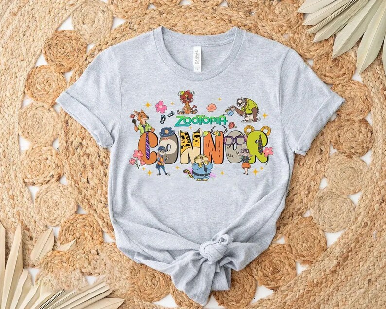 Puede incluir: Camiseta gris claro con la palabra "Connor" en letras coloridas estilo caricatura, y la palabra "Zootopia" arriba. El dise&ntilde;o incluye varios personajes de dibujos animados y acentos florales, creando una est&eacute;tica juguetona y vibrante.