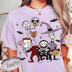 May include: Lilac Comfort Colors t-shirt featuring characters from The Nightmare Before Christmas. The design includes Jack Skellington, Oogie Boogie, and other characters, along with Mickey Mouse-shaped balloons. The shirt has the text "Comfort Color Orchid".