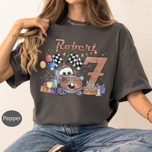 May include: A charcoal gray t-shirt featuring a cartoon illustration of Mater from the movie Cars, with the name "Robert" and the number "7". The design includes checkered flags, balloons, and a birthday cake, celebrating a seventh birthday.