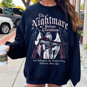May include: Black sweatshirt featuring the text "The Nightmare Before Christmas" above an illustration of Jack Skellington, the Pumpkin King. The graphic includes a figure holding a book with red accents and the text "Halloween Town 1993".