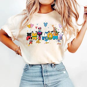 May include: Cream-coloured t-shirt with the words "Mickey & Friends" in colourful, cartoon-style lettering. Mickey Mouse, Minnie Mouse, Donald Duck, and other Disney characters are incorporated into the design, with balloons and hearts.