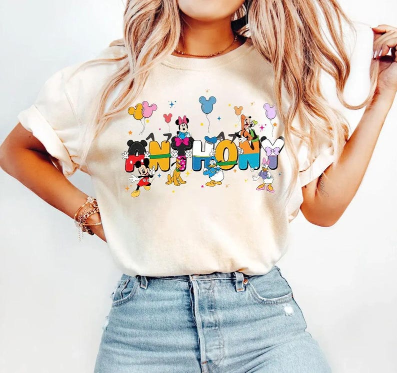 May include: Cream-coloured t-shirt with the words "Mickey & Friends" in colourful, cartoon-style lettering. Mickey Mouse, Minnie Mouse, Donald Duck, and other Disney characters are incorporated into the design, with balloons and hearts.