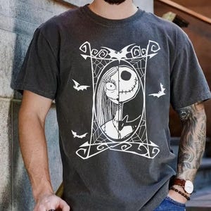 May include: Dark gray t-shirt featuring a white graphic of Jack Skellington and Sally from The Nightmare Before Christmas. The design is framed with a web and bat silhouettes. The shirt has short sleeves and a crew neck.