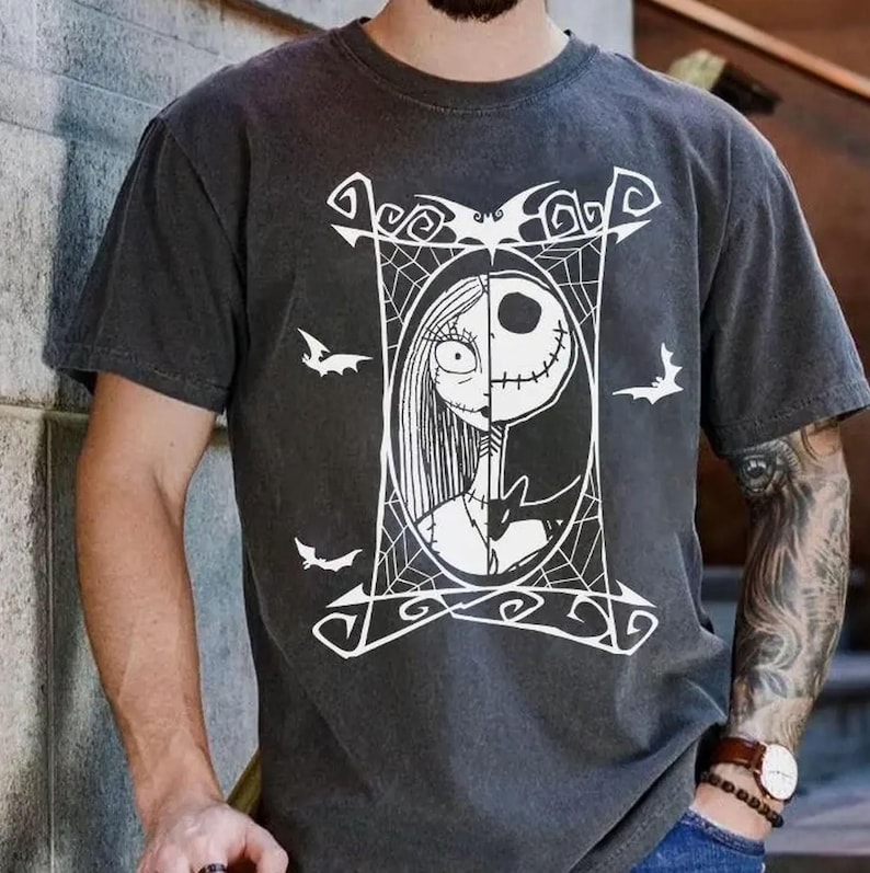 May include: Dark gray t-shirt featuring a white graphic of Jack Skellington and Sally from The Nightmare Before Christmas. The design is framed with a web and bat silhouettes. The shirt has short sleeves and a crew neck.