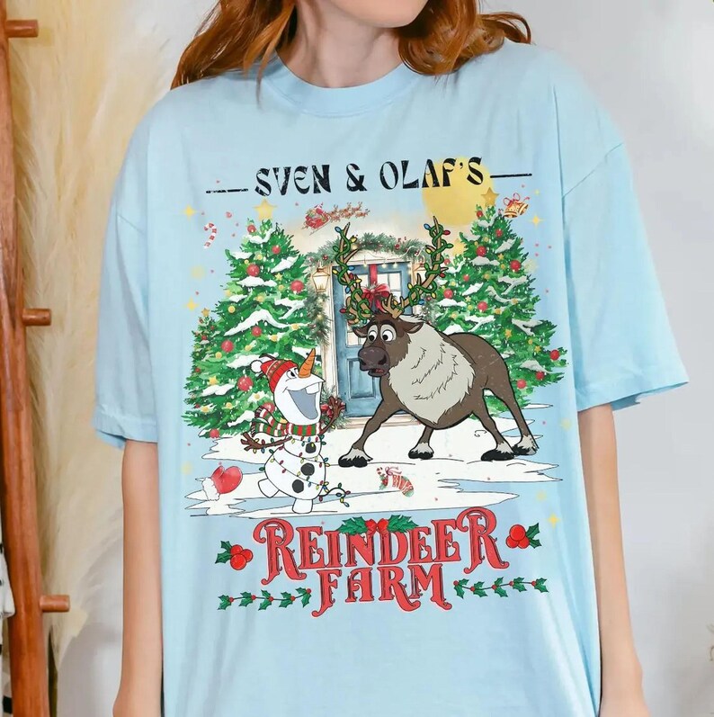 May include: Light blue t-shirt featuring a festive design with Sven the reindeer and Olaf the snowman. The shirt has the text "Sven & Olaf's" above a winter scene with the words "Reindeer Farm" below.
