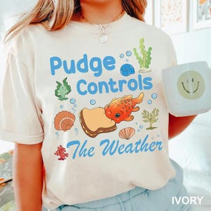 May include: Ivory-colored t-shirt with the text "Pudge Controls The Weather" in blue. The design includes an orange fish, a sandwich, seashells, and seaweed. A white mug with a smiley face is held in the image.