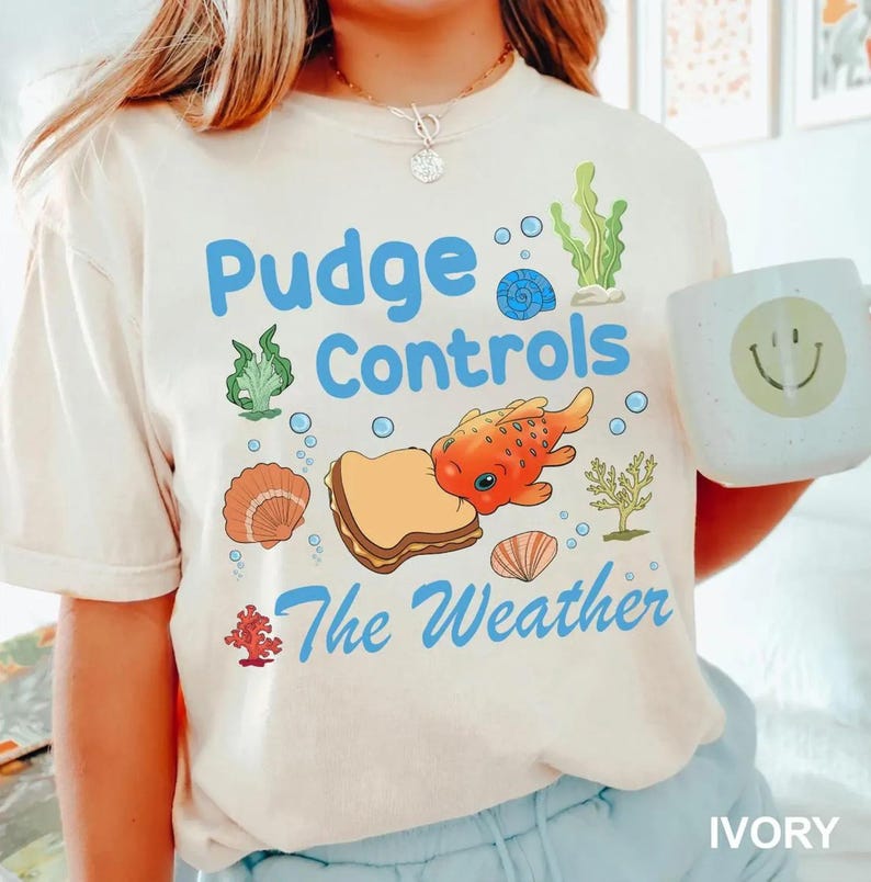 May include: Ivory-colored t-shirt with the text "Pudge Controls The Weather" in blue. The design includes an orange fish, a sandwich, seashells, and seaweed. A white mug with a smiley face is held in the image.