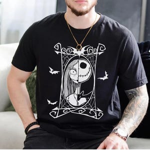May include: Black t-shirt featuring a white graphic of Jack Skellington and Sally from The Nightmare Before Christmas. The design is framed by a decorative border with bats and spiderwebs. A gold chain necklace is visible.