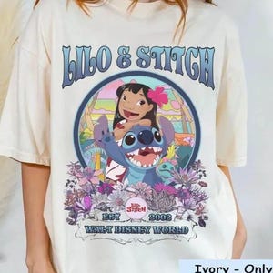 Może przedstawiać: Kremowa koszulka z okrągłą grafiką Lilo i Stitch. Projekt zawiera tekst "Lilo & Stitch", "EST 2002" i "Walt Disney World". Grafika przedstawia Lilo z kwiatem we włosach i Stitcha. Koszulka jest Comfort Color.