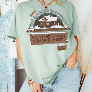 Op de afbeelding: Lichtgroen T-shirt met een afbeelding van "Big Summer Blowout" en een hutillustratie. Het shirt toont ook "Wandering Oaken's Trading Post" en somt zomerartikelen op.