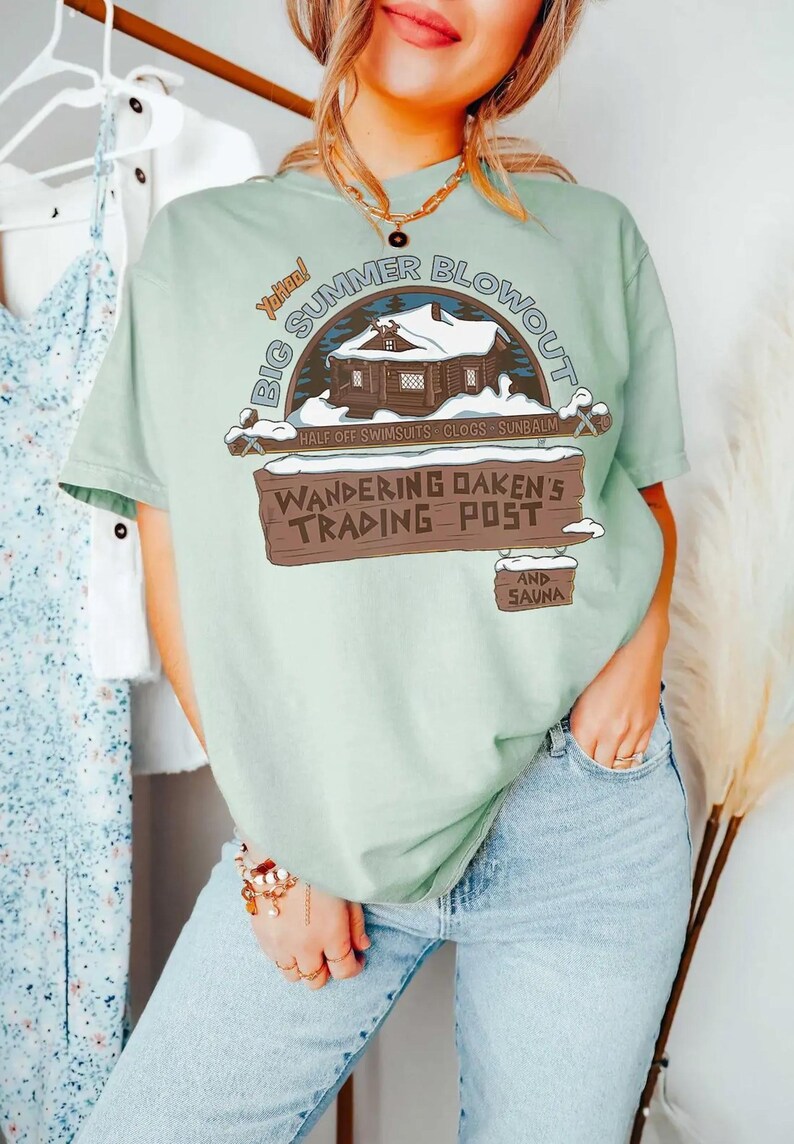 Op de afbeelding: Lichtgroen T-shirt met een afbeelding van "Big Summer Blowout" en een hutillustratie. Het shirt toont ook "Wandering Oaken's Trading Post" en somt zomerartikelen op.