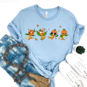 May include: Light blue t-shirt featuring four cartoon orange characters with green leaves and orange details. The characters are in different poses, with flowers and stars around them. The shirt is paired with blue jeans and white sneakers.