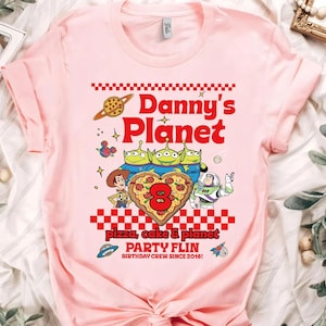 Pu&ograve; includere: T-shirt rosa chiaro con un design grafico colorato. Il design include il testo "Danny's Planet" in rosso, insieme a personaggi dei cartoni animati e le parole "pizza, cake & planet" e "PARTY FLIN BIRTHDAY CREW SINCE 2016!"