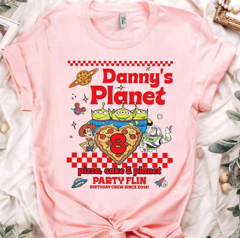 Pu&ograve; includere: T-shirt rosa chiaro con un design grafico colorato. Il design include il testo "Danny's Planet" in rosso, insieme a personaggi dei cartoni animati e le parole "pizza, cake & planet" e "PARTY FLIN BIRTHDAY CREW SINCE 2016!"