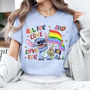 May include: Light blue t-shirt featuring a cartoon character holding a rainbow flag. The shirt has the text "ALL FOR LOVE AND LOVE FOR ALL" in colorful letters. The design includes stars, hearts, and a small bear.
