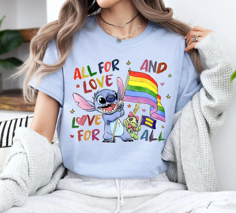 May include: Light blue t-shirt featuring a cartoon character holding a rainbow flag. The shirt has the text "ALL FOR LOVE AND LOVE FOR ALL" in colorful letters. The design includes stars, hearts, and a small bear.