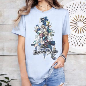 Puede incluir: Camiseta azul claro con un gr&aacute;fico colorido de personajes de Kingdom Hearts, incluyendo a Mickey Mouse y Donald Duck. El dise&ntilde;o presenta varios personajes de la serie de videojuegos. El texto "Kingdom Hearts" est&aacute; impreso debajo.
