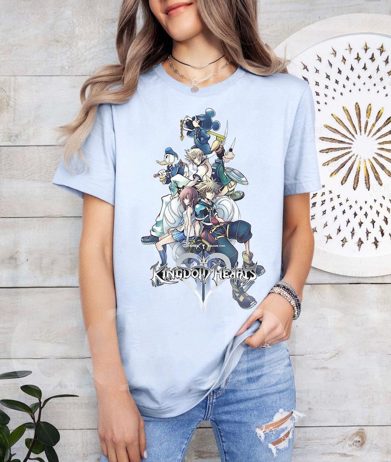 Puede incluir: Camiseta azul claro con un gr&aacute;fico colorido de personajes de Kingdom Hearts, incluyendo a Mickey Mouse y Donald Duck. El dise&ntilde;o presenta varios personajes de la serie de videojuegos. El texto "Kingdom Hearts" est&aacute; impreso debajo.