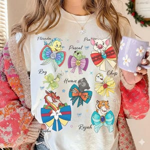 Puede incluir: Camiseta color crema con personajes de dibujos animados con lazos coloridos y nombres como Flounder, Pascal y Pua. El diseño incluye mariposas y estrellas. Es una prenda de vestir informal.
