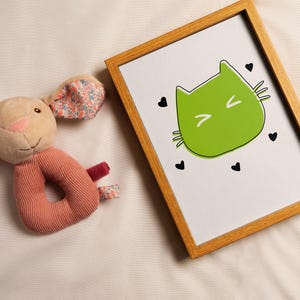 May include: A framed print featuring a green cat face with a black outline and black heart accents. A plush bunny rattle with floral patterned ears and a ribbed pink body is next to the frame. The background is a light beige fabric.
