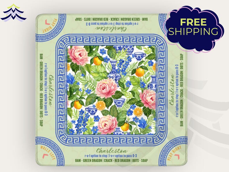 May include: A square decorative item with a floral design featuring pink roses, blue flowers, and oranges. The item has a blue and white border with the word "Charleston" and other text. The item is on a light green background.