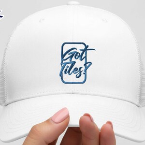 Op de afbeelding: Witte truckerpet met een mesh achterkant en een gebogen klep. De voorkant van de pet heeft de geborduurde tekst "Got Tiles?" in blauw schrift binnen een blauwe rechthoekige rand. De pet wordt door een persoon vastgehouden.