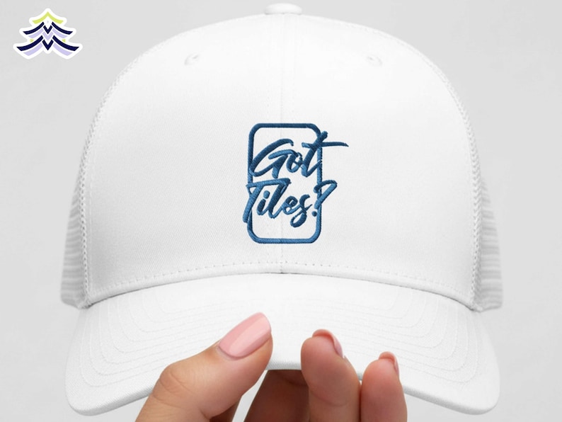 Op de afbeelding: Witte truckerpet met een mesh achterkant en een gebogen klep. De voorkant van de pet heeft de geborduurde tekst "Got Tiles?" in blauw schrift binnen een blauwe rechthoekige rand. De pet wordt door een persoon vastgehouden.