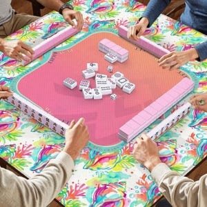 以下が含まれることがあります： ピンク色のゲーム面とカラフルな模様の縁取りがある正方形の麻雀卓。テーブルの周りにはプレイヤーがいて、それぞれピンク色の牌立てを持っています。白い麻雀牌とサイコロがテーブルに散らばっています。