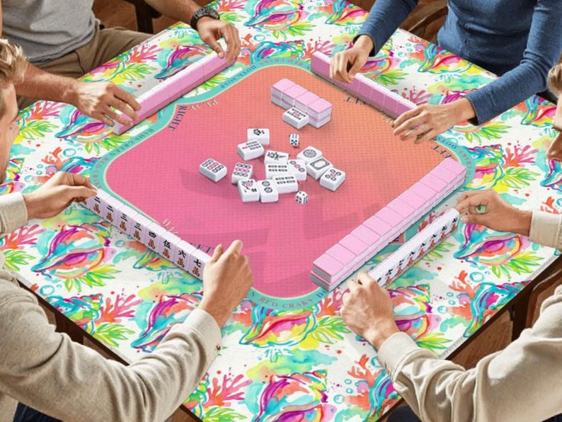 以下が含まれることがあります： ピンク色のゲーム面とカラフルな模様の縁取りがある正方形の麻雀卓。テーブルの周りにはプレイヤーがいて、それぞれピンク色の牌立てを持っています。白い麻雀牌とサイコロがテーブルに散らばっています。