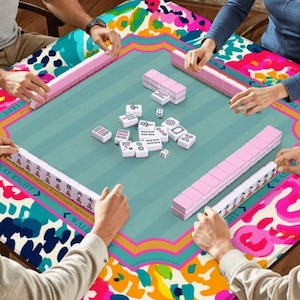 May include: A group of people playing Mahjong on a square table. The table has a teal center with pink and white Mahjong tiles, surrounded by a colorful floral pattern. The players are holding pink tile racks. The table's edge has the text "PLAY RIGHT".