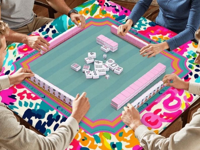 May include: A group of people playing Mahjong on a square table. The table has a teal center with pink and white Mahjong tiles, surrounded by a colorful floral pattern. The players are holding pink tile racks. The table's edge has the text "PLAY RIGHT".