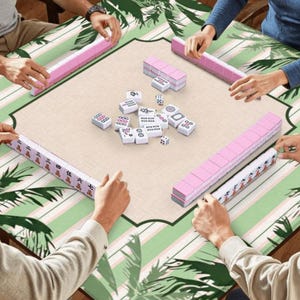 K&ouml;nnte beinhalten: Eine Mahjong-Partie, bei der Spieler um einen Tisch sitzen. Der Tisch hat ein gr&uuml;nes und wei&szlig;es tropisches Bl&auml;ttermuster. Rosa Kachelregale und Kacheln sind angeordnet, zusammen mit W&uuml;rfeln. Die Spieler halten die Kacheln.